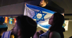 Vīrietis tur Izraēlas karogu, kamēr cilvēki skatās 2025. gada Eirovīzijas dziesmu konkursa finālu.