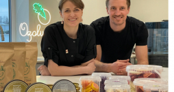 Marta un Intars Ozoli savā jaunajā ražotnē pie lielā darba galda, uz kura izlikts teju viss viņu ražojumu klāsts, kā arī “Novada garšas” konkursā iegūtās godalgas. Priekšplānā – gabaliņos sagrieztā medus kūka.