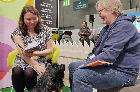 Bērnu grāmatu autore Emilija Tolvanena (no kreisās) un sunīte klausītāja Hloja ar instruktori Helsinku grāmatu izstādes klausītājsuņu stendā. Bērnu grāmatu autore Emilija Tolvanena (no kreisās) un sunīte klausītāja Hloja ar instruktori Helsinku grāmatu izstādes klausītājsuņu stendā.