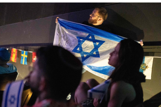 Vīrietis tur Izraēlas karogu, kamēr cilvēki skatās 2025. gada Eirovīzijas dziesmu konkursa finālu.