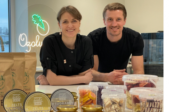 Marta un Intars Ozoli savā jaunajā ražotnē pie lielā darba galda, uz kura izlikts teju viss viņu ražojumu klāsts, kā arī “Novada garšas” konkursā iegūtās godalgas. Priekšplānā – gabaliņos sagrieztā medus kūka.