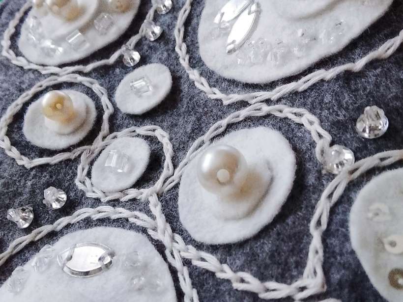 Tuvplānā redzama visa izšūto pērļu un fliteru bagātība un baltās dzijas izšūtās “taciņas”.