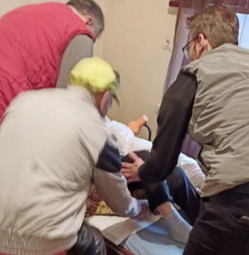 Kliente no auto līdz mājas durvīm tika atvesta ar transportēšanas gultu. Taču viņu neieveda mājās ar gultu vai ratiņkrēslā, bet nesa pa betona pakāpieniem, aiz segas stūriem turēdami. Istabā uzņemtajā attēlā apguldīšanas brīdis. Redzams, ka nav ne transportēšanas gultas, ne ratiņkrēsla. Citās reizēs mājās tika braukts gan ar gultu, gan ratiņkrēslu.