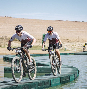 Kārlis Krūmiņš (priekšplānā) un Ādolfs Žunna slavenajā "Cape Epic" kalnu riteņbraukšanas maratonā pa improvizētu tiltiņu pievārē kādu ūdensšķērsli.