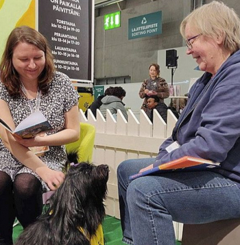 Bērnu grāmatu autore Emilija Tolvanena (no kreisās) un sunīte klausītāja Hloja ar instruktori Helsinku grāmatu izstādes klausītājsuņu stendā.