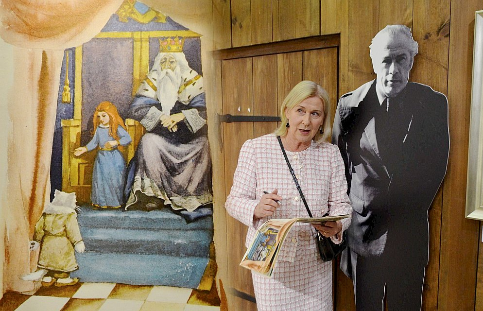 Mākslinieka meita Ilze Zvagūze-Parsons pie tēva foto un mākslinieka ilustrācijas K. Skalbes pasakai “Kaķīša dzirnavām”.