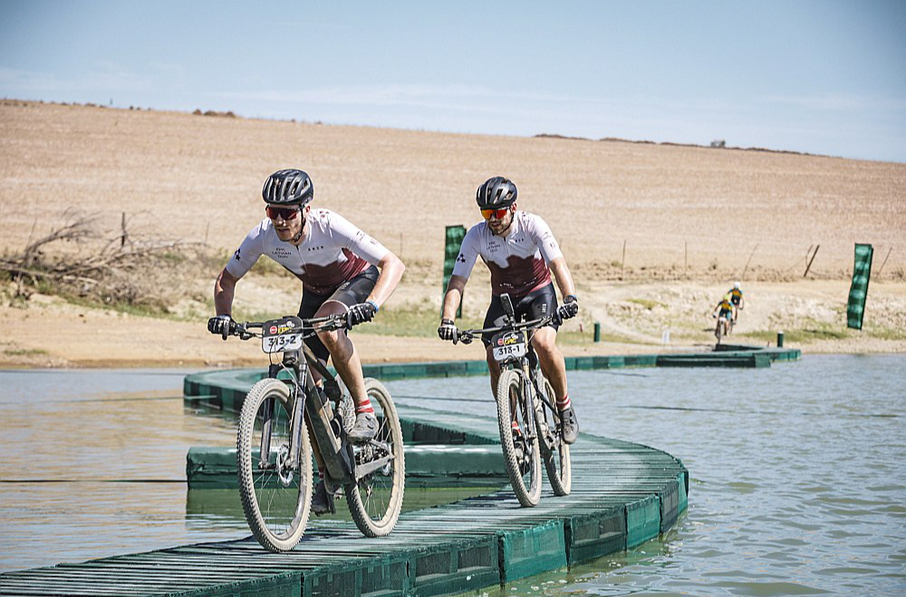 Kārlis Krūmiņš (priekšplānā) un Ādolfs Žunna slavenajā "Cape Epic" kalnu riteņbraukšanas maratonā pa improvizētu tiltiņu pievārē kādu ūdensšķērsli.