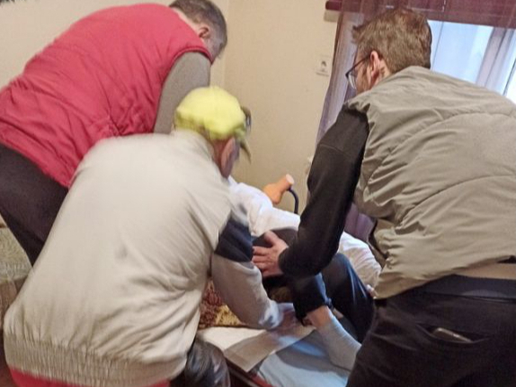 Kliente no auto līdz mājas durvīm tika atvesta ar transportēšanas gultu. Taču viņu neieveda mājās ar gultu vai ratiņkrēslā, bet nesa pa betona pakāpieniem, aiz segas stūriem turēdami. Istabā uzņemtajā attēlā apguldīšanas brīdis. Redzams, ka nav ne transportēšanas gultas, ne ratiņkrēsla. Citās reizēs mājās tika braukts gan ar gultu, gan ratiņkrēslu.