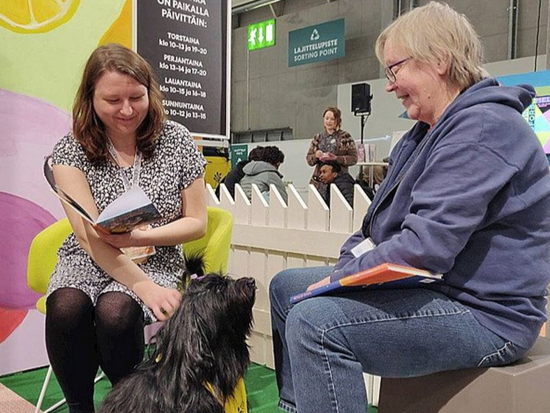 Bērnu grāmatu autore Emilija Tolvanena (no kreisās) un sunīte klausītāja Hloja ar instruktori Helsinku grāmatu izstādes klausītājsuņu stendā.