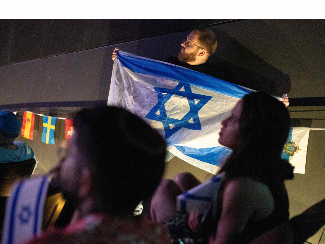 Vīrietis tur Izraēlas karogu, kamēr cilvēki skatās 2025. gada Eirovīzijas dziesmu konkursa finālu.