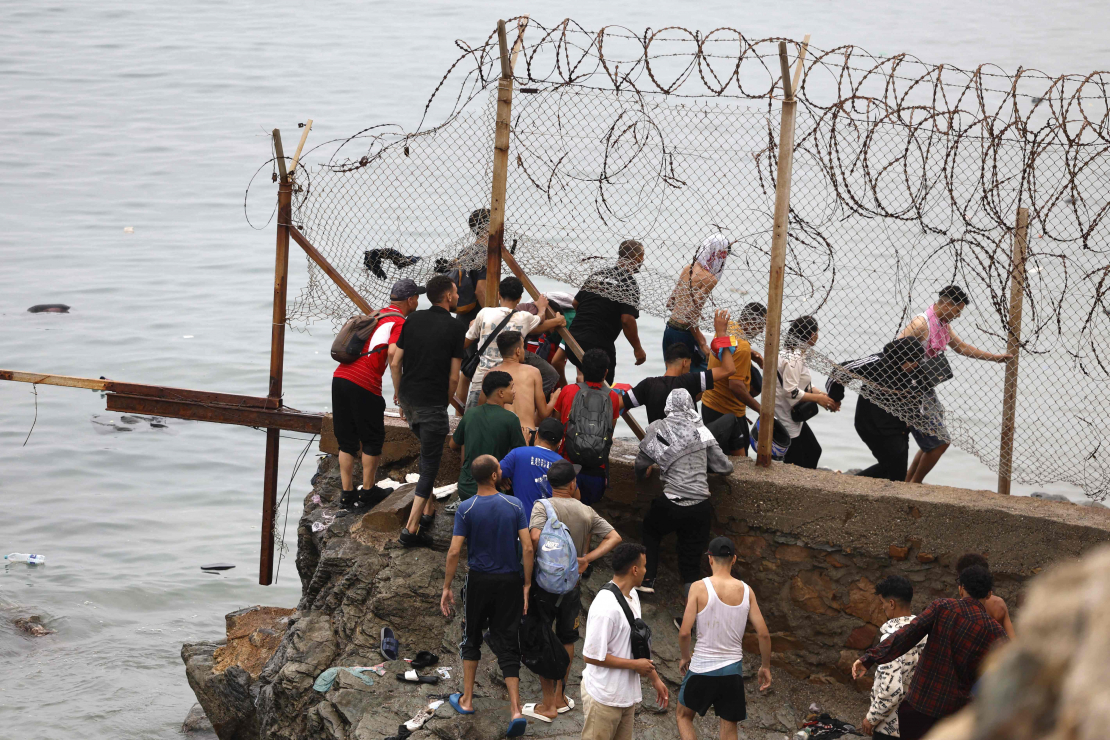 Pie žoga netālu no sadursmju vietas pie Fnidekas, uz Marokas un Spānijas robežas, 2026. gada 31. jūlijā pulcējušies cilvēki. Tūkstošiem migrantu nakts laikā no Marokas šķērsoja robežu un iekļuva Spānijas Ziemeļāfrikas anklāvā Seutā, 31. jūlijā pavēstīja policijas avots.