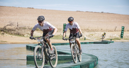 Kārlis Krūmiņš (priekšplānā) un Ādolfs Žunna slavenajā "Cape Epic" kalnu riteņbraukšanas maratonā pa improvizētu tiltiņu pievārē kādu ūdensšķērsli.
