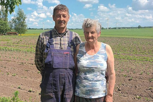 Gunārs un Laimute Kātiņi, stāvot savā piemājas dārziņā, iezīmē "robu" fona ainavā, kur 15 gadus stāvēja aprīļa beigās nogāzusies vēja elektrostacija. "Vairāk esam uztraukti par tiem torņiem, kuri ir daudz tuvāk pie mājas un joprojām griežas," viņi saka.