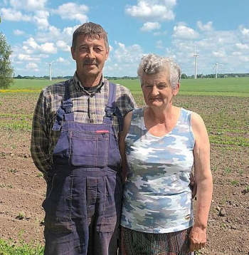Gunārs un Laimute Kātiņi, stāvot savā piemājas dārziņā, iezīmē "robu" fona ainavā, kur 15 gadus stāvēja aprīļa beigās nogāzusies vēja elektrostacija. "Vairāk esam uztraukti par tiem torņiem, kuri ir daudz tuvāk pie mājas un joprojām griežas," viņi saka.