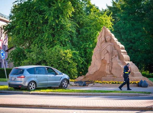 Sabojāta smilšu skulptūras apakšējā daļa, bet automobilim sabojāts priekšējais buferis un dzinēja pārsega sāni.
