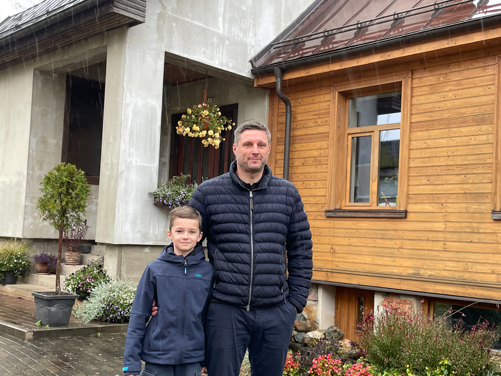 Saimnieks Ivars Grantiņš ar dēlu Ernestu mājas pagalmā, kur labi redzams, kā senākā daļa savienota ar no jauna uzbūvēto nodegušā apjoma
vietā.