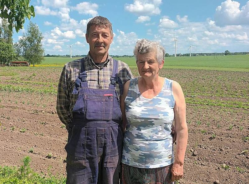 Gunārs un Laimute Kātiņi, stāvot savā piemājas dārziņā, iezīmē "robu" fona ainavā, kur 15 gadus stāvēja aprīļa beigās nogāzusies vēja elektrostacija. "Vairāk esam uztraukti par tiem torņiem, kuri ir daudz tuvāk pie mājas un joprojām griežas," viņi saka.
